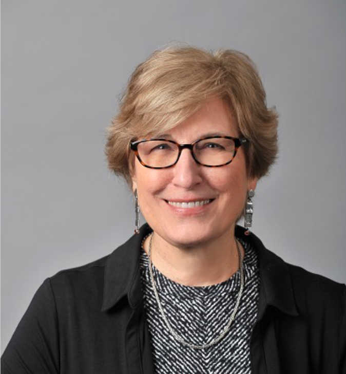 Headshot of a School of Social Work faculty member with short blonde hair and glasses, smiling, wearing a black blazer over a patterned top and long earrings against a neutral gray background.
